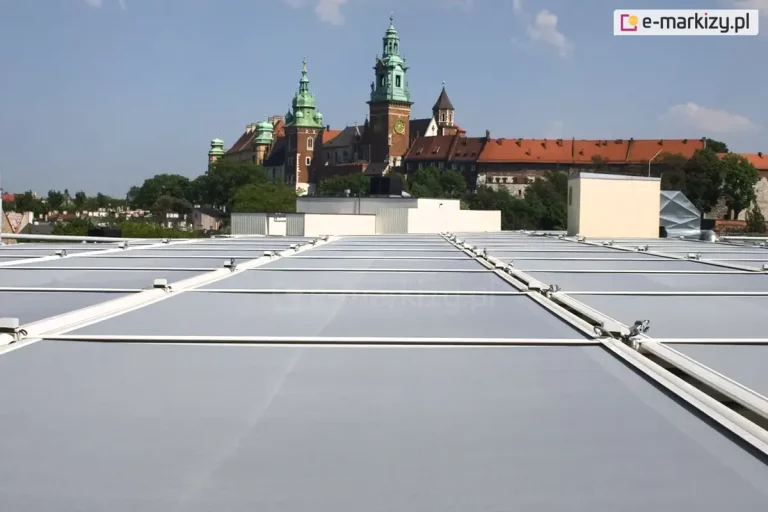 Wielomodułowy system veranda zamontowany na płaskim dachu budynku, widoczny z perspektywy górnej. Jasnoszara tkanina akrylowa jest rozpięta w kilku równoległych modułach oddzielonych białymi prowadnicami aluminiowymi. Pomiędzy kasetami modułów zainstalowano puszki hermetyczne do zasilania napędów. Markizy tworzą jednolitą osłonę przeciwsłoneczną nad dużą powierzchnią przeszkloną. W tle, za krawędzią dachu, znajduje się panorama Krakowa z wyraźnie widocznym Zamkiem Królewskim na Wawelu, wieżami katedry oraz murami obronnymi pod pogodnym, błękitnym niebem.