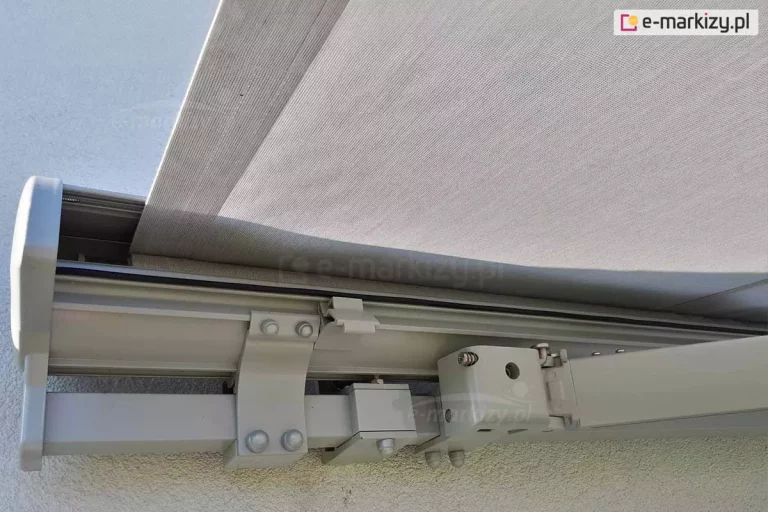 Techniczne zbliżenie na detale konstrukcyjne oraz mocowanie markizy tarasowej Palladio w kolorze jasnoszarym, którą zawieszono na ścianie o jasnej elewacji budynku. Widok od dołu ukazuje profil nośny o kwadratowym przekroju, do którego za pomocą masywnych, uchwytów przymocowana jest kaseta. Elementy mocujące są skręcone stalowymi śrubami z widocznymi łbami w kolorze konstrukcji. Po prawej stronie kadru znajduje się mechanizm przegubowy ramienia markizy, który odpowiada za jej rozkładanie i stabilizację. Pomiędzy tymi elementami widoczny jest uchwyt mocujący markizę do ściany zabezpieczony od dołu śrubą z nakrętką i zabezpieczony zaślepką. Poszycie markizy wykonane jest z gęstej tkaniny akrylowej o drobnoziarnistym splocie w kolorze szarym, która wychodzi z górnej części kasety. Całość konstrukcji jest utrzymana w nowoczesnej, minimalistycznej estetyce, a precyzyjne wykończenie lakiernicze profili aluminiowych oraz solidne punkty styku komponentów świadczą o wysokiej jakości wykonania systemu osłonowego.