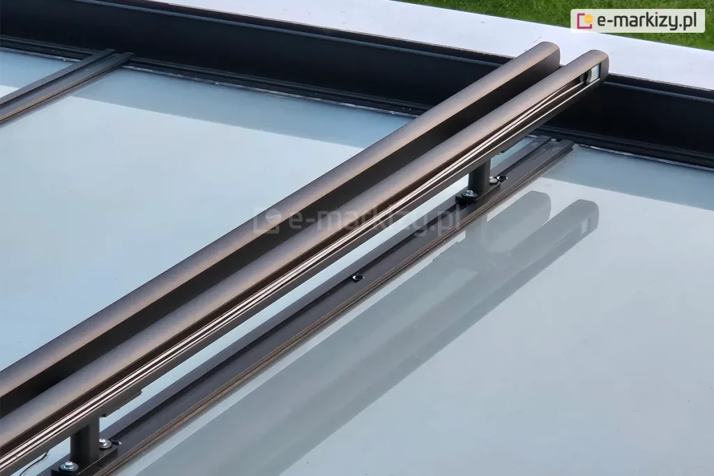 Zdjęcie przedstawia szczegółowe ujęcie konstrukcji prowadnic modułowych systemu veranda, zamontowanego nad szklanym dachem. Dwie równoległe, aluminiowe prowadnice w kolorze ciemnego antracytu o matowym wykończeniu biegną ukośnie przez kadr, wspierając się na pionowych wspornikach dystansowych. Wsporniki te są przykręcone do krokwi zadaszenia za pomocą stalowych śrub, a cała konstrukcja unosi się kilka centymetrów nad taflą szkła. W czystej, szklanej powierzchni dachu wyraźnie odbijają się elementy metalowego stelaża oraz niebo, tworząc lustrzany efekt. Po prawej stronie, na krawędzi konstrukcji, widoczne są elementy skrzynek osłon przednich, za którymi rozpościera się zielony trawnik ogrodu.