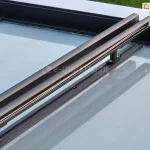 Zdjęcie przedstawia szczegółowe ujęcie konstrukcji prowadnic modułowych systemu veranda, zamontowanego nad szklanym dachem. Dwie równoległe, aluminiowe prowadnice w kolorze ciemnego antracytu o matowym wykończeniu biegną ukośnie przez kadr, wspierając się na pionowych wspornikach dystansowych. Wsporniki te są przykręcone do krokwi zadaszenia za pomocą stalowych śrub, a cała konstrukcja unosi się kilka centymetrów nad taflą szkła. W czystej, szklanej powierzchni dachu wyraźnie odbijają się elementy metalowego stelaża oraz niebo, tworząc lustrzany efekt. Po prawej stronie, na krawędzi konstrukcji, widoczne są elementy skrzynek osłon przednich, za którymi rozpościera się zielony trawnik ogrodu.