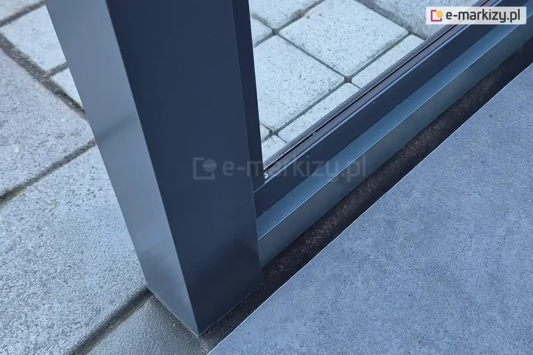 Mocowanie stałej ściany szklanej Fest do zadaszenia Lunaro. Pomiędzy podporami zainstalowane zostało okno w ramie. Pod zadaszeniem widoczny taras wyłożony betonowymi płytkami i kostką.