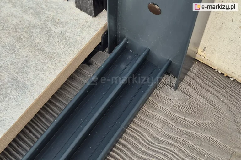 Dolna prowadnica systemu szyb przesuwnych Miros w kolorze antracytowym zamocowana na drewnianym tarasie w pobliżu profilu bocznego doszczelniającego U także w kolorze antracytowym. Po lewej stronie trzytorowej prowadnicy widoczna gruba płyta gresowa w jasnym kolorze.