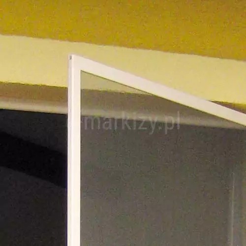 Ramka moskitiery ramkowej na zawiasach Soft w kolorze białym z szarą siatką widoczna na tle okna i żółtej ściany.