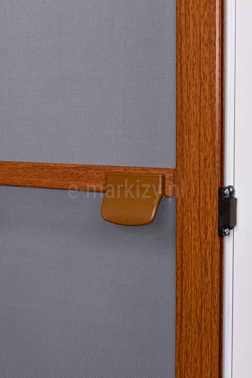 Zbliżenie elementów moskitiery Saloon - klamka w kolorze brązowym zamocowana do szprosu moskitiery Saloon w drewnopodobnym kolorze. Obok widoczny czarny magnes, który umożliwia skuteczne domknięcie drzwi moskitierowych do ramy okna.