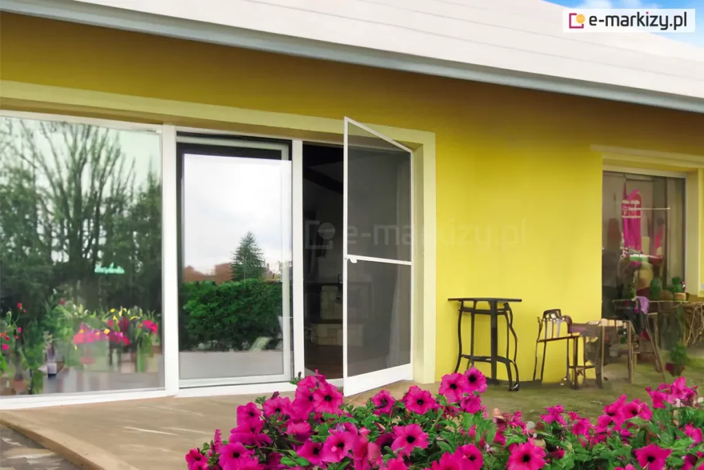 Moskitiera drzwiowa na zawiasach Soft zamocowana do ramy okna o przesuwnym skrzydle z białą ramą. Lekko uchylona siatka pozwala dostrzec we wnętrzu piec kaflowy, a biała rama kontrastuje z żółtą ścianą elewacji, przy której ustawiono różne przedmioty o dziwnych kształtach. Całość widoczna z perspektywy kwietnika pełnego różowo kwitnących surfinii.