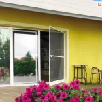 Moskitiera drzwiowa na zawiasach Soft zamocowana do ramy okna o przesuwnym skrzydle z białą ramą. Lekko uchylona siatka pozwala dostrzec we wnętrzu piec kaflowy, a biała rama kontrastuje z żółtą ścianą elewacji, przy której ustawiono różne przedmioty o dziwnych kształtach. Całość widoczna z perspektywy kwietnika pełnego różowo kwitnących surfinii.