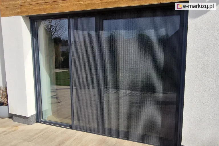 Szerokie okno tarasowe w ścianie o jasnej elewacji, nad którym znajduje się ozdobny drewniany panel, przesłonięte zostały częściowo otwartą moskitierą plisowaną Zig Zag zamocowaną do wnęki okiennej przy tarasie z dużych płyt w kolorze jasnego drewna.