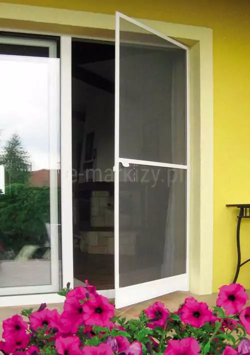 Moskitiera ramkowa na zawiasach Soft w kolorze białym zamocowana do ramy okna ze skrzydłem przesuwnym o białym kolorze. Okno umieszczone we wnęce o białej ramie, w ścianie o żółtym kolorze. Przed wyjściem na taras widoczne różowe kwiaty surfinii.