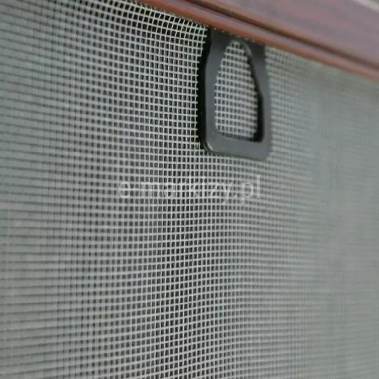 Zbliżenie czarnego uchwytu do podtrzymania moskitiery ramkowej podczas montażu na oknie, wsuniętego pomiędzy profil aluminiowy kołnierza moskitiery w kolorze brązowym a szarą siatkę.