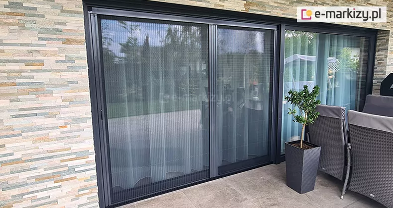 Duże okno tarasowe w ścianie wyłożonej wąskimi płytkami w odcieniach brązu i szarości osłonięte zostało podwójną moskitierą plisowaną Click Rol. Zapewnia ona skuteczną osłonę przed insektami. Po prawej stronie przed oknem na tarasie widoczne krzesła ratanowe z pokrowcami oraz wysoka czarna kwadratowa doniczka z niskim drzewkiem.