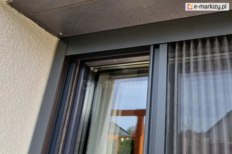Lekko otwarta szara siatka moskitiery plisowanej Top Zag o konstrukcji w kolorze antracytowym, zamontowanej do wnęki okiennej okna o antracytowej ramie wyposażonego także w rolety zewnętrzne. Profile boczne moskitiery przymocowane tuż przy prowadnicy rolety.