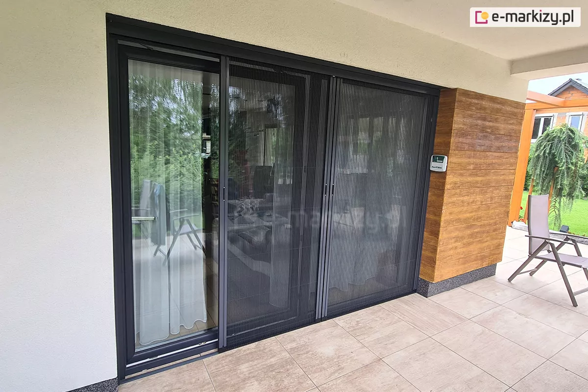 Niewielki taras w narożniku domu, z wyjściem przez duże okno z przesuwnym skrzydłem w antracytowej ramie. Okno zostało zasłonięte moskitierą plisowaną z czarną siatką i antracytową konstrukcją złożoną z dwóch paneli. Jeden z modułów siatki lekko odsunięty ukazuje okno przesłonięte białą firaną do samej podłogi. Na tarasie wyłożonym dużymi płytkami w kolorze beżowo-szarym, przy którym ściany wyłożono częściowo drewnianymi listwami, a częściowo wykończono białym tynkiem, stoi krzesło z tkaninowym siedziskiem i oparciem. W tle widoczny ogród oraz jedno drzewko iglaste.