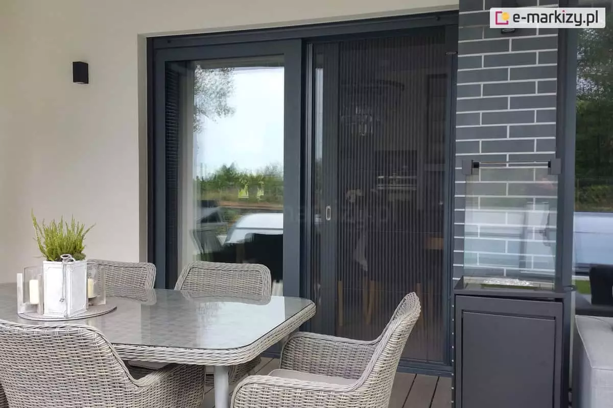 Czarne drzwi tarasowe HS osłonięte moskitierą plisowaną Zig Zag z czarną siatką, która chroni przed komarami wnętrze pomieszczenia. Przed drzwiami ustawiono wiklinowy stół nakryty szybą z ustawionym na niej kwietnikiem i lampionami. Wokół stołu ustawione cztery dopasowane wiklinowe krzesła. Obok, po prawej stronie widoczny wygaszony kominek na biopaliwo.