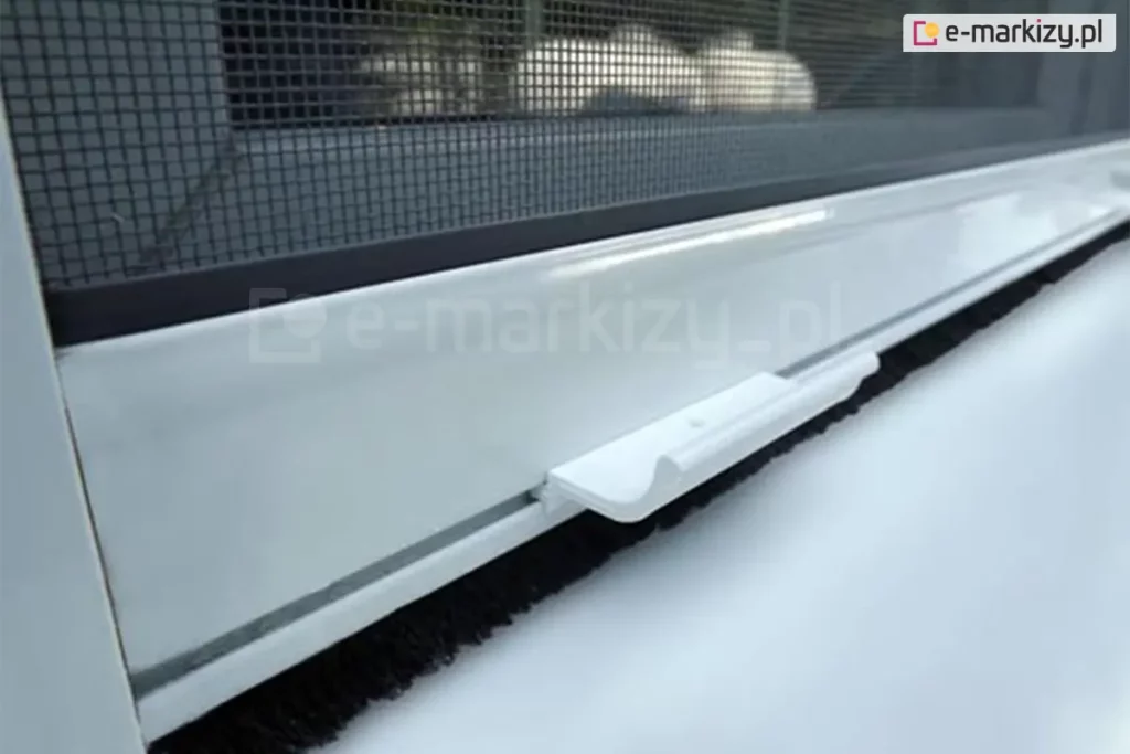 Zbliżenie białej belki dolnej moskitiery okiennej rolowanej z widocznym mocowaniem siatki, klamką ułatwiającą otwieranie i zamykanie oraz uszczelką szczotkową, zabezpieczającą przed owadami na poziomie parapetu okna.
