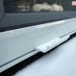 Zbliżenie białej belki dolnej moskitiery okiennej rolowanej z widocznym mocowaniem siatki, klamką ułatwiającą otwieranie i zamykanie oraz uszczelką szczotkową, zabezpieczającą przed owadami na poziomie parapetu okna.