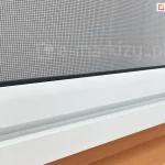 Zbliżenie belki dolnej z siatką przeciw komarom zaczepionej o prowadnicę dolną moskitiery rolowanej do okna dachowego lub tradycyjnego widoczne nad drewnianym parapetem okiennym.