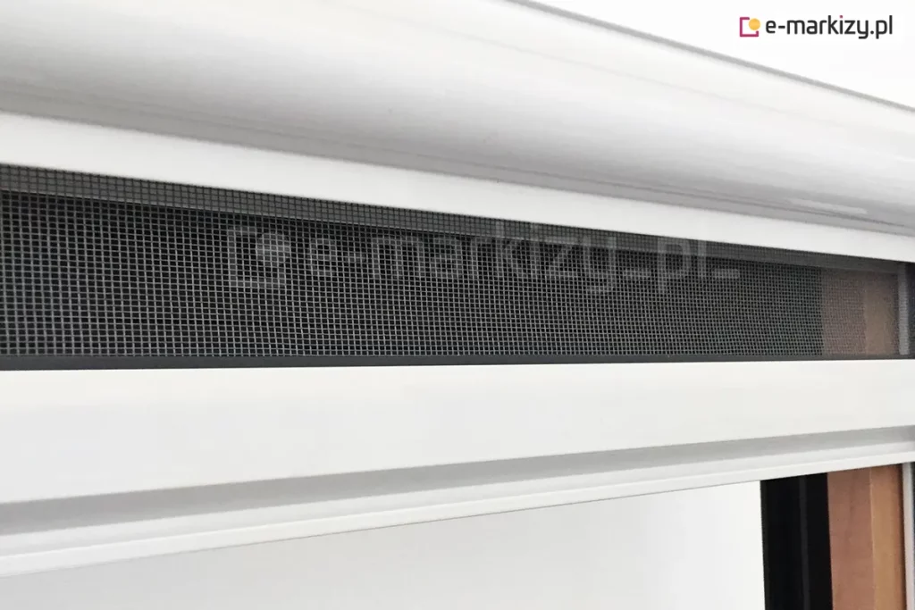 Zbliżenie kasety oraz częściowo wysuniętej siatki szarej zamocowanej w belce dolnej moskitiery rolowanej z zaczepem hakowym, stosowanym do okien dachowych. Moskitiera z aluminiową kasetą, prowadnicami i belką wykonaną w kolorze białym zamocowana na oknie o drewnianej framudze.