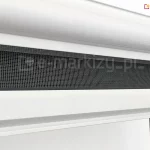 Zbliżenie kasety oraz częściowo wysuniętej siatki szarej zamocowanej w belce dolnej moskitiery rolowanej z zaczepem hakowym, stosowanym do okien dachowych. Moskitiera z aluminiową kasetą, prowadnicami i belką wykonaną w kolorze białym zamocowana na oknie o drewnianej framudze.