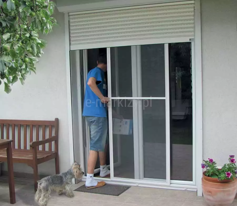 Moskitiera przesuwna Slim w białym kolorze zamocowana pomiędzy ramą okna a białą roletą zewnętrzną, przesunięta przez wchodzącego do pomieszczenia mężczyznę w krótkich dżinsowych spodenkach, niebieskiej koszulce, białych skarpetkach i kapciach, trzymającego w jednej ręce ulotkę. Za mężczyzną stojącym jedną nogą na wycieraczce przed wejściem czeka pies ray terrier. Na tarasie widoczna także drewniana ławka i stół oraz ustawiona po przeciwnej stronie okna donica z petuniami o biało-filetowych kwiatach.