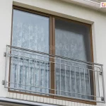 Moskitiera przesuwna Solid z jednym panelem z siatką umieszczona na brązowym oknie balkonowym z francuskim balkonem. Nad oknem widoczna podbitka dachu w białym kolorze. Okno dodatkowo od środka pomieszczenia przesłonięto firanką w różany wzór.