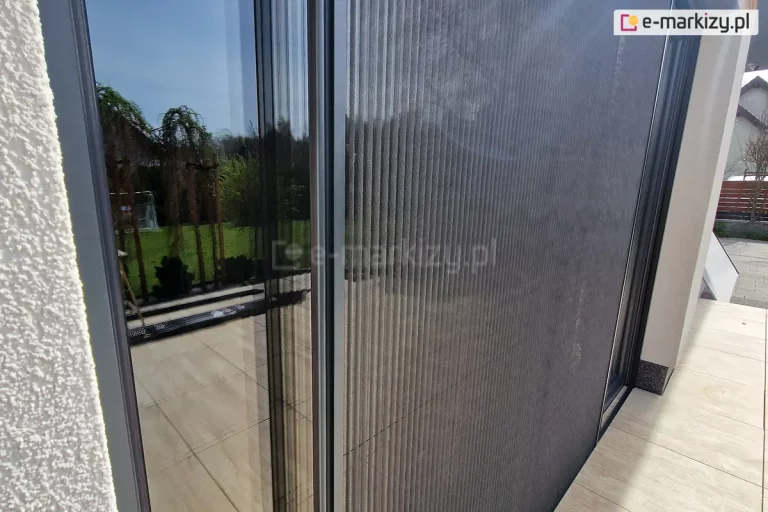 Wnęka okienna o białym tynku strukturalnym z oknem tarasowym w odcieniach antracytu i dopasowaną kolorystycznie moskitierą plisowaną Top Zag, o dwustronnym otwieraniu.