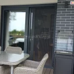 Model moskitiery plisowanej na drzwi Top Zag wykonany w kolorystyce grafitowej, dopasowany do koloru ramy okiennej, w której został zamocowany. Na tarasie przed oknem widoczny stół przykryty na blacie taflą szkła z dopasowanymi do niego krzesłami.