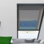 Moskitiera rolowana na okno dachowe w czarnej ramie, z częściowo opuszczoną czarną siatką, zamontowana w jasnym wnętrzu poddasza. Przez siatkę widać widok na las, obok znajduje się sofa z poduszkami i nowoczesna lampka ścienna.