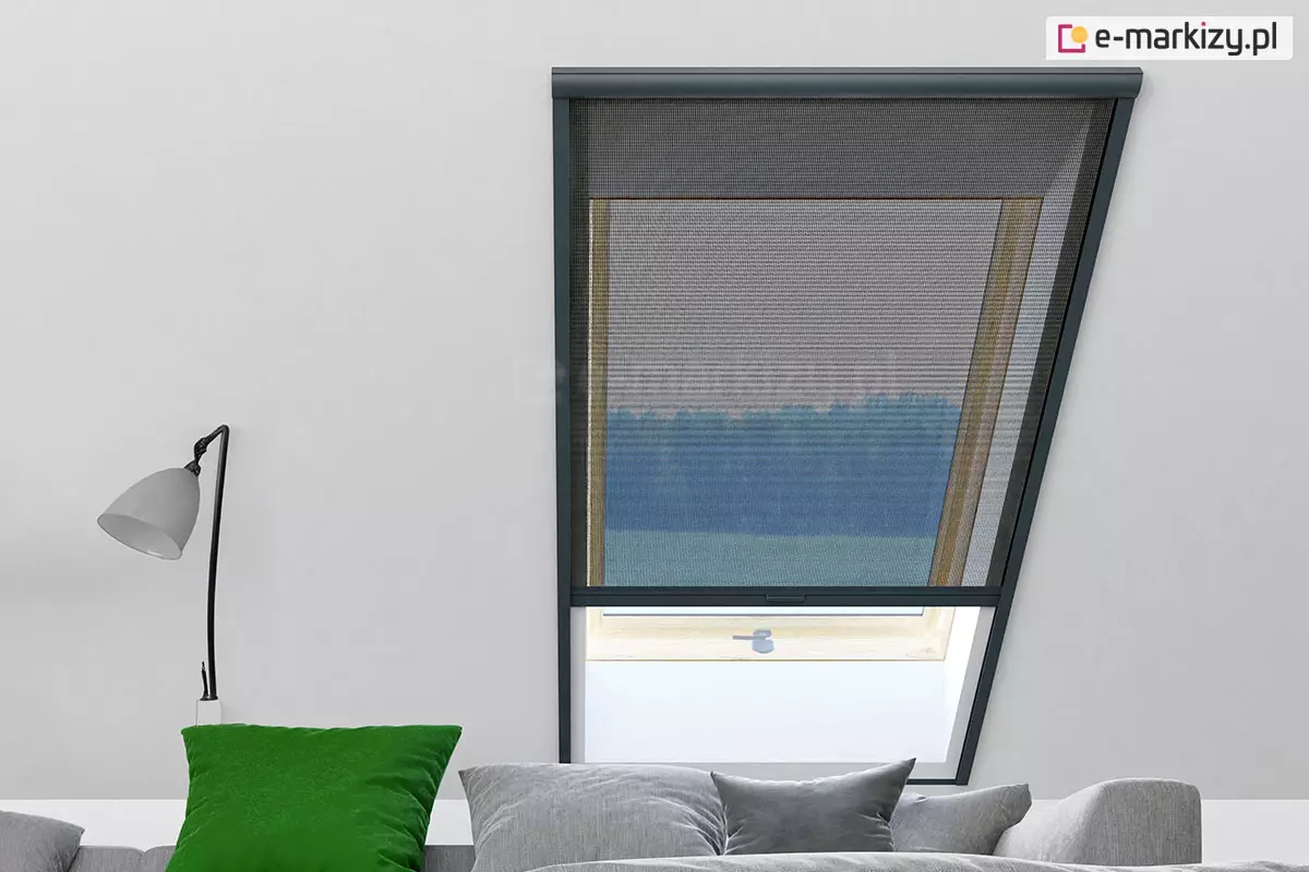 Moskitiera rolowana na okno dachowe w czarnej ramie, z częściowo opuszczoną czarną siatką, zamontowana w jasnym wnętrzu poddasza. Przez siatkę widać widok na las, obok znajduje się sofa z poduszkami i nowoczesna lampka ścienna.