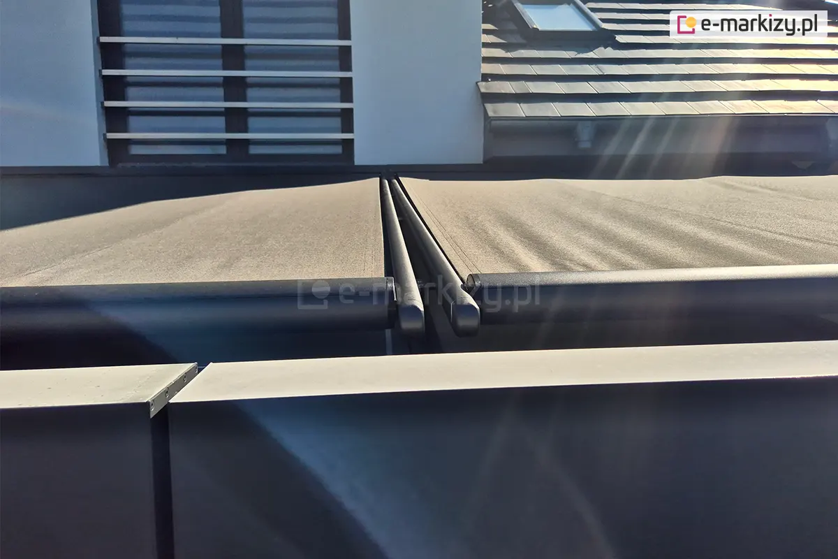 Zbliżenie z perspektywy górnej na detal wykończenia i prowadnice dwóch sąsiadujących ze sobą rolet veranda w kolorze antracytowym. Na pierwszym planie widoczne są dwa zaokrąglone, metalowe profile obciążające, które stanowią zakończenie rozwijanego materiału; profile te mają gładką, matową powierzchnię i są zakończone estetycznymi zaślepkami z tworzywa. Poszycie markiz wykonane z tkaniny o wyraźnym splocie jest mocno napięte, a jego powierzchnia delikatnie mieni się w intensywnym świetle słonecznym, tworząc subtelne smugi i refleksy. Wąska szczelina między dwoma niezależnymi modułami markiz ukazuje precyzję montażu systemów dwuczęściowych. W tle widoczny jest fragment elewacji budynku z jasnym tynkiem oraz fragment dachu pokrytego nowoczesną, płaską dachówką w kolorze grafitowym z wbudowanym oknem połaciowym. Po lewej stronie znajduje się wysokie okno z zamontowaną zewnętrzną barierką francuską składającą się z poziomych, metalowych szczebli. Silne nasłonecznienie z prawej strony kadru rzuca głębokie cienie i podkreśla teksturę materiałów oraz geometryczne linie całej konstrukcji zadaszenia.
