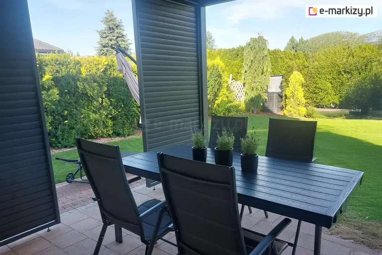 Zdjęcie przedstawia widok z wnętrza nowoczesnej pergoli tarasowej na przydomowy ogród. Na pierwszym planie znajduje się zestaw mebli ogrodowych w kolorze czarnym lub ciemnoantracytowym, składający się z dużego, prostokątnego stołu o blacie z szerokich listew oraz pasujących do niego krzeseł z wysokimi oparciami. Na stole ustawione są trzy czarne, okrągłe doniczki z zielonymi ziołami, prawdopodobnie lawendą. Konstrukcja pergoli wyposażona jest w panele boczne wypełnione poziomymi, szerokimi lamelami w tym samym ciemnym kolorze co meble. Jeden z paneli jest ustawiony centralnie, dzieląc widok na ogród na dwie części, natomiast drugi widoczny jest po lewej stronie kadru. Podłoże tarasu wyłożone jest prostokątną kostką brukową w odcieniach brązu i szarości. W tle rozciąga się zadbany trawnik oraz gęste nasadzenia z tuj, iglaków i krzewów ozdobnych w różnych odcieniach zieleni i żółci, które tworzą szczelną barierę roślinną. Po lewej stronie, za panelami, widoczny jest fragment złożonego parasola ogrodowego w kolorze beżowym oraz mały wózek ogrodowy. Całość sceny oświetlona jest naturalnym, dziennym światłem, co podkreśla kontrast między ciemną zabudową a jaskrawą zielenią ogrodu.