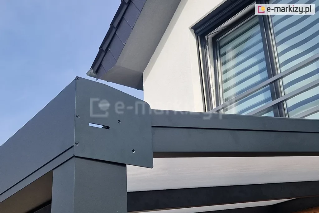 Zbliżenie na zewnętrzne połączenie narożne zadaszenia tarasowego Modes, gdzie stykają się belki poziome oraz pionowy słup nośny. Punkt ten został zabezpieczony płaską, antracytową maskownicą o nieregularnym kształcie, przykręconą za pomocą czterech czarnych wkrętów. Element ten estetycznie maskuje zakończenie rynny oraz krawędź bocznego wypełnienia dachu, które w tym przypadku stanowi biała, nieprzezierna płyta. W tle widoczna jest biała elewacja piętra budynku z oknem wyposażonym w nowoczesne rolety zewnętrzne oraz barierka francuska.