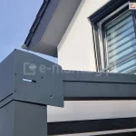 Zbliżenie na zewnętrzne połączenie narożne zadaszenia tarasowego Modes, gdzie stykają się belki poziome oraz pionowy słup nośny. Punkt ten został zabezpieczony płaską, antracytową maskownicą o nieregularnym kształcie, przykręconą za pomocą czterech czarnych wkrętów. Element ten estetycznie maskuje zakończenie rynny oraz krawędź bocznego wypełnienia dachu, które w tym przypadku stanowi biała, nieprzezierna płyta. W tle widoczna jest biała elewacja piętra budynku z oknem wyposażonym w nowoczesne rolety zewnętrzne oraz barierka francuska.