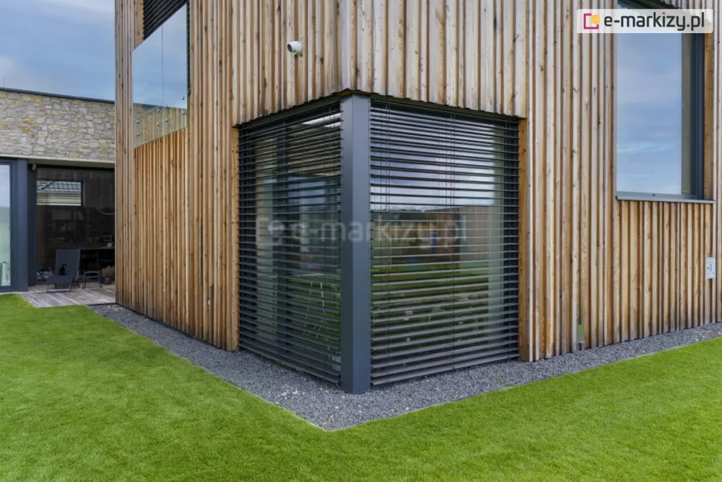Narożne żaluzje fasadowe Z90 Box firmy Selt w kolorze antracytowym zamontowane zostały na fasadzie domku wykończonej drewnianymi panelami. Przed oknem widoczna otoczka z szarych kamieni oraz zadbany trawnik. W tle widoczny kolejny budynek parterowy.