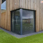 Narożne żaluzje fasadowe Z90 Box firmy Selt w kolorze antracytowym zamontowane zostały na fasadzie domku wykończonej drewnianymi panelami. Przed oknem widoczna otoczka z szarych kamieni oraz zadbany trawnik. W tle widoczny kolejny budynek parterowy.