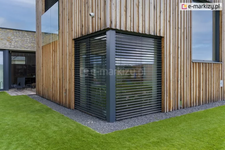 Narożne żaluzje fasadowe Z90 Box firmy Selt w kolorze antracytowym zamontowane zostały na fasadzie domku wykończonej drewnianymi panelami. Przed oknem widoczna otoczka z szarych kamieni oraz zadbany trawnik. W tle widoczny kolejny budynek parterowy.