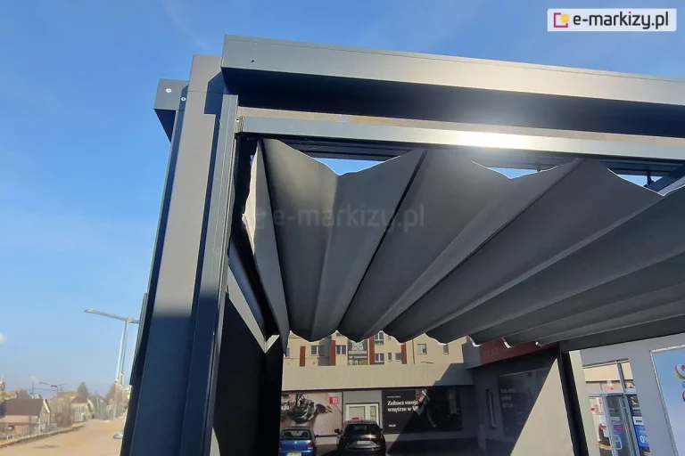 Narożnik konstrukcji pergoli wolnostojącej tako w kolorze grafitowym z rozwiniętym dachem rzymskim i widocznymi refleksolami od boku i frontu. Pod pergolą widoczny budynek i auta na parkingu. Po lewej stronie ulica z domkami i błękitne niebo.
