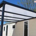 Zdjęcie prezentuje detal konstrukcyjny aluminiowego zadaszenia tarasowego Nebbiolo marki Deponti, zamontowanego do ściany jasnego domku o elewacji z poziomych paneli. Perspektywa z dołu uwidacznia szkielet dachu wykonany z solidnych profili aluminiowych w kolorze antracytowym, składający się z belki przyściennej, krokwi oraz rynny frontowej zintegrowanej z narożnym łącznikiem. Wypełnienie dachu stanowią płyty poliwęglanu komorowego o mlecznej, opalowej barwie, które równomiernie rozpraszają światło. Całość konstrukcji wspiera się na pionowym słupie o kwadratowym przekroju, widocznym po lewej stronie kadru. W tle, pod zadaszeniem, widoczne są białe framugi drzwi i okien budynku, a powyżej linia drzew liściastych na tle bezchmurnego, błękitnego nieba.