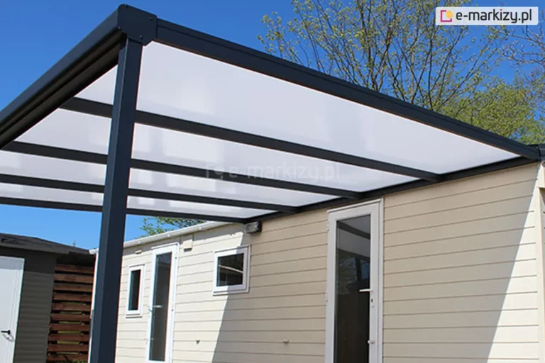 Zdjęcie prezentuje detal konstrukcyjny aluminiowego zadaszenia tarasowego Nebbiolo marki Deponti, zamontowanego do ściany jasnego domku o elewacji z poziomych paneli. Perspektywa z dołu uwidacznia szkielet dachu wykonany z solidnych profili aluminiowych w kolorze antracytowym, składający się z belki przyściennej, krokwi oraz rynny frontowej zintegrowanej z narożnym łącznikiem. Wypełnienie dachu stanowią płyty poliwęglanu komorowego o mlecznej, opalowej barwie, które równomiernie rozpraszają światło. Całość konstrukcji wspiera się na pionowym słupie o kwadratowym przekroju, widocznym po lewej stronie kadru. W tle, pod zadaszeniem, widoczne są białe framugi drzwi i okien budynku, a powyżej linia drzew liściastych na tle bezchmurnego, błękitnego nieba.