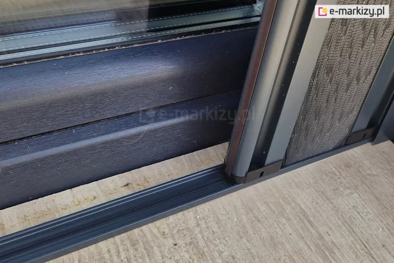 Bardzo niski - zaledwie 8 mm wysokości próg dolny moskitiery plisowanej Top Zag z widocznymi w jego wnętrzu sznureczkami naklejony na powierzchnię płytek tarasowych z drewnianym wzorem. Na progu umieszczony pakiet z maksymalnie zwiniętą siatką. Próg umieszczony w oddaleniu od ramy okna ze względu na rolety zewnętrzne.