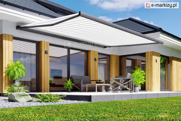 Zdjęcie przedstawia nowoczesny dom jednorodzinny o parterowej bryle z rozległym tarasem, nad którym zamontowana jest duża, wysuwana markiza tarasowa Moderno MOL. Markiza posiada białe poszycie o delikatnej strukturze materiału, które jest rozpięte na solidnym stelażu w kolorze ciemnoszarym lub antracytowym. Konstrukcja wspierająca markizę składa się z bocznych ramion przegubowych zamontowanych w kasecie zawieszonej bezpośrednio do elewacji budynku. Taras jest wyłożony jasnoszarymi płytami o dużym formacie, a jego krawędź wykończona jest ciemniejszymi bloczkami betonowymi. Na tarasie znajduje się zestaw mebli ogrodowych w nowoczesnym stylu, składający się z ciemnoszarej sofy, fotela z dekoracyjnymi, krzyżującymi się elementami konstrukcyjnymi oraz niskiego stolika kawowego o drewnianym blacie. Przestrzeń wypoczynkowa jest udekorowana roślinami doniczkowymi w ciemnych i szarych donicach, w tym palmami oraz mniejszymi paprociami. Po lewej stronie zdjęcia widoczna jest aranżacja ogrodowa z dekoracyjnym głazem i drobnym, szarym żwirem. Elewacja domu łączy biały tynk z pionowymi panelami wykonanymi z jasnego drewna, które znajdują się pomiędzy dużymi, przeszklonymi drzwiami tarasowymi w ciemnych ramach. Dach budynku jest pokryty ciemną dachówką płaską, a na jego połaci widoczne są panele fotowoltaiczne. W tle rozciąga się zadbany trawnik oraz błękitne niebo z lekkimi, białymi chmurami. Cała kompozycja jest jasna, słoneczna i prezentuje estetyczne połączenie architektury z funkcjonalnym rozwiązaniem osłonowym.