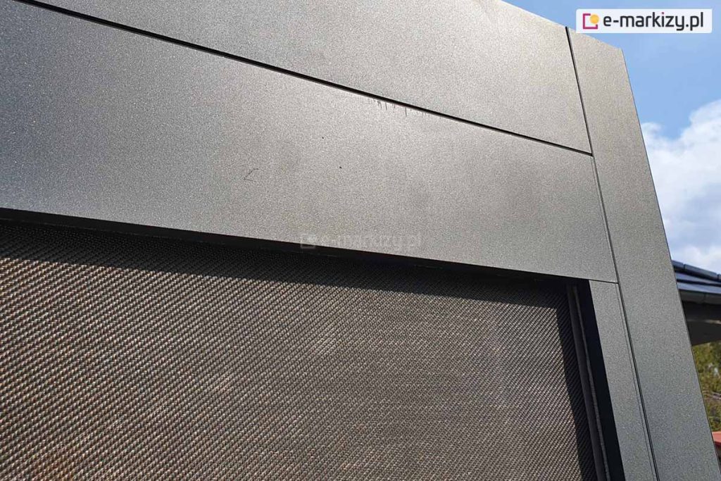 Zdjęcie prezentuje szczegółowe zbliżenie na górny narożnik nowoczesnej pergoli aluminiowej Luxury, skupiając się na estetyce wykończenia i strukturze materiałów. Na pierwszym planie widoczne są solidne, szerokie profile aluminiowe w kolorze ciemnografitowym o matowej, drobnoziarnistej strukturze, połączone pod kątem prostym. Pod poziomą belką konstrukcyjną zamontowana jest dopasowana kolorystycznie kaseta, z której rozwinięta jest tkanina techniczna typu screen w kolorze brązowo-szarym o gęstym, diagonalnym splocie. Materiał ten jest precyzyjnie osadzony w prowadnicy bocznej zintegrowanej z pionowym słupem pergoli. W tle, po prawej stronie, dostrzec można fragment krawędzi dachu budynku oraz jasne, lekko zachmurzone niebo. Ujęcie podkreśla wysoką jakość spasowania elementów metalowych oraz techniczną precyzję wykonania bocznych osłon przeciwsłonecznych.