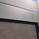 Zdjęcie prezentuje szczegółowe zbliżenie na górny narożnik nowoczesnej pergoli aluminiowej Luxury, skupiając się na estetyce wykończenia i strukturze materiałów. Na pierwszym planie widoczne są solidne, szerokie profile aluminiowe w kolorze ciemnografitowym o matowej, drobnoziarnistej strukturze, połączone pod kątem prostym. Pod poziomą belką konstrukcyjną zamontowana jest dopasowana kolorystycznie kaseta, z której rozwinięta jest tkanina techniczna typu screen w kolorze brązowo-szarym o gęstym, diagonalnym splocie. Materiał ten jest precyzyjnie osadzony w prowadnicy bocznej zintegrowanej z pionowym słupem pergoli. W tle, po prawej stronie, dostrzec można fragment krawędzi dachu budynku oraz jasne, lekko zachmurzone niebo. Ujęcie podkreśla wysoką jakość spasowania elementów metalowych oraz techniczną precyzję wykonania bocznych osłon przeciwsłonecznych.