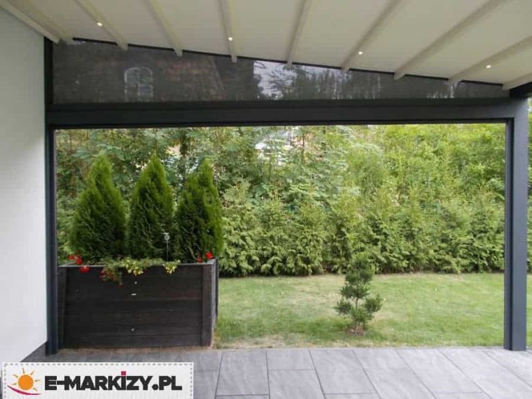 Widok z wnętrza tarasu na nowoczesną pergolę elektryczną Moderno w ciemnej kolorystyce, wyposażoną w biały, segmentowy dach tkaninowy z zamontowanym oświetleniem LED w formie punktowych diod na profilach poprzecznych. Pod konstrukcją dachu, na ścianie bocznej, zamontowano trójkątny panel wypełniający z ciemnej, transparentnej tkaniny technicznej typu screen. Na tarasie wyłożonym szarymi płytami znajduje się duża, czarna donica drewniana z trzema uformowanymi krzewami iglastymi i drobnymi kwiatami, a w tle rozpościera się widok na ogród z gęstym żywopłotem i zadbanym trawnikiem.