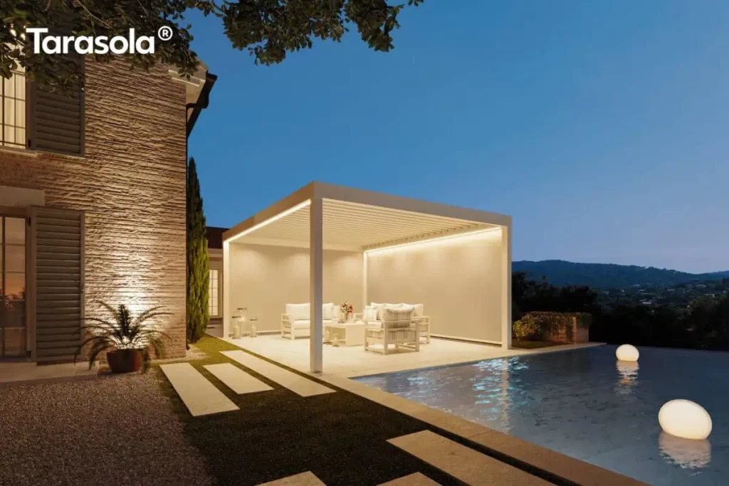 Nowoczesna pergola lamelowa essential Tarasola w kolorze białym lub kremowym, zaprezentowana w scenerii wieczornej z włączonym oświetleniem LED zintegrowanym wzdłuż ramy dachu. Wolnostojąca konstrukcja bioklimatyczna z dachem z ruchomych lameli oświetla wnętrze strefy wypoczynkowej, w której znajduje się komplet białych mebli tarasowych z jasnymi poduszkami. Pergola została posadowiona na tarasie z jasnych płyt kamiennych bezpośrednio przy basenie typu infinity, w którym pływają dwie świecące kule dekoracyjne. Obok znajduje się ściana budynku z jasnej, nieregularnej cegły z szarymi okiennicami, oświetlona punktowym światłem podkreślającym jej fakturę. Otoczenie obejmuje precyzyjnie przycięty trawnik z wkomponowanymi kamiennymi płytami chodnikowymi, palmę w donicy oraz wysoki, smukły iglak. W tle, pod granatowym, wieczornym niebem, widoczna jest panorama wzgórz i świateł miejscowości, co tworzy nastrojową i elegancką atmosferę luksusowej przestrzeni rekreacyjnej.
