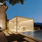 Nowoczesna pergola lamelowa essential Tarasola w kolorze białym lub kremowym, zaprezentowana w scenerii wieczornej z włączonym oświetleniem LED zintegrowanym wzdłuż ramy dachu. Wolnostojąca konstrukcja bioklimatyczna z dachem z ruchomych lameli oświetla wnętrze strefy wypoczynkowej, w której znajduje się komplet białych mebli tarasowych z jasnymi poduszkami. Pergola została posadowiona na tarasie z jasnych płyt kamiennych bezpośrednio przy basenie typu infinity, w którym pływają dwie świecące kule dekoracyjne. Obok znajduje się ściana budynku z jasnej, nieregularnej cegły z szarymi okiennicami, oświetlona punktowym światłem podkreślającym jej fakturę. Otoczenie obejmuje precyzyjnie przycięty trawnik z wkomponowanymi kamiennymi płytami chodnikowymi, palmę w donicy oraz wysoki, smukły iglak. W tle, pod granatowym, wieczornym niebem, widoczna jest panorama wzgórz i świateł miejscowości, co tworzy nastrojową i elegancką atmosferę luksusowej przestrzeni rekreacyjnej.