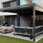 Fragment nowoczesnego ogrodu z tarasem, którego głównym elementem jest minimalistyczne zadaszenie Square Flat wykonane z ciemnoszarego metalu. Konstrukcja składa się z prostych słupów i dachu tkaninowego z belkami przeciwwietrznymi, w których zainstalowano oświetlenie LED. Pod pergolą ustawiona jest narożna sofa ogrodowa na drewnianej podstawie, z siedziskami i oparciami w kolorze jasnoszarym, osadzonymi w ciemnej ramie. Po lewej stronie, na jednym z segmentów sofy, leży żółta poduszka. Tło stanowi nowoczesna elewacja domu w dwóch kontrastujących odcieniach – bieli i antracytu – z dużymi oknami tarasowymi zasłoniętymi poziomymi żaluzjami. Na lewym skraju zdjęcia widoczna jest strefa relaksu z dwoma leżakami ogrodowymi ustawionymi na płytach tarasowych, obok grilla i niskiej donicy. Całą przestrzeń otaczają starannie utrzymane nasadzenia – smukłe drzewa iglaste, brzozy oraz żywopłot przy grafitowym ogrodzeniu.