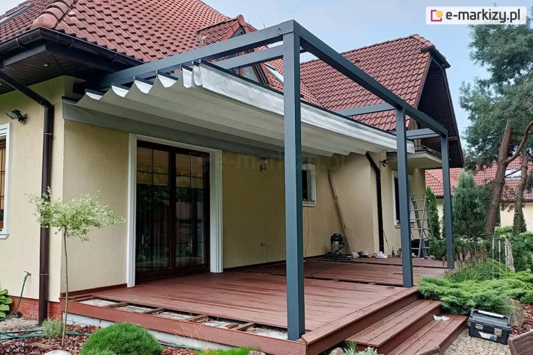 Aluminiowa pergola tarasowa Tako w kolorze antracytowym z zadaszeniem rzymskim, zamontowana do elewacji domu jednorodzinnego z dachem krytym czerwoną dachówką. Jasna tkanina zadaszenia jest częściowo rozwinięta, tworząc dekoracyjne fałdy podwieszone pod konstrukcją. Pergola wspiera się na trzech pionowych słupach osadzonych na drewnianym tarasie z brązowych desek, do którego prowadzą szerokie schody. Pod zadaszeniem widoczne są duże, przeszklone drzwi tarasowe w brązowych ramach oraz jasna elewacja budynku. Wokół tarasu znajduje się ogród z niskimi krzewami iglastymi, trawą dekoracyjną oraz wysokimi sosnami w tle. Na tarasie widoczne są przedmioty wskazujące na trwające prace montażowe: odkurzacz przemysłowy, drabina aluminiowa oraz skrzynka narzędziowa.