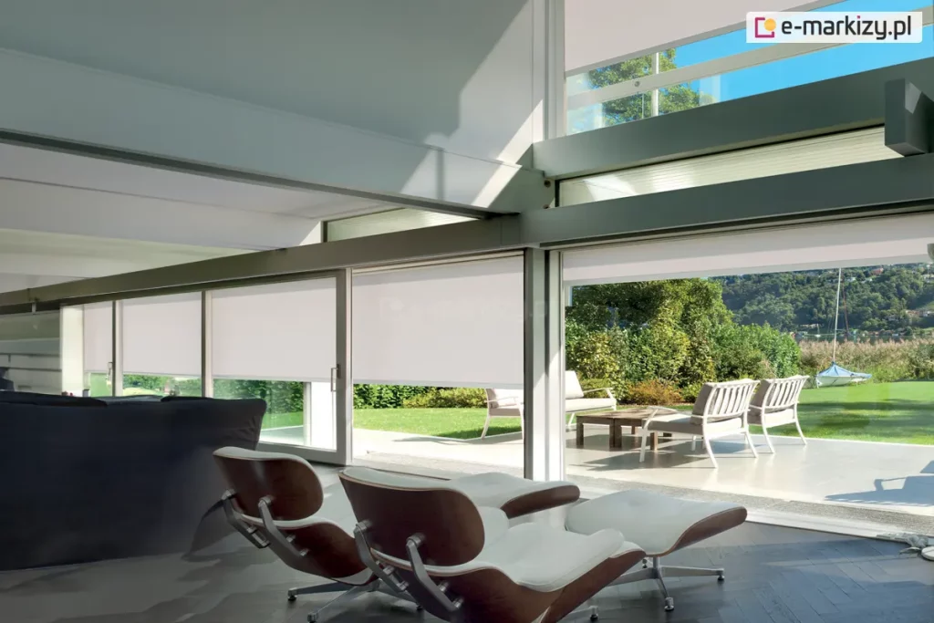 Nowoczesne rolety zaciemniające z półprzezroczystą tkaniną w jasnoszarym odcieniu i widokiem na taras i ogród