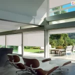 Nowoczesne rolety zaciemniające z półprzezroczystą tkaniną w jasnoszarym odcieniu i widokiem na taras i ogród