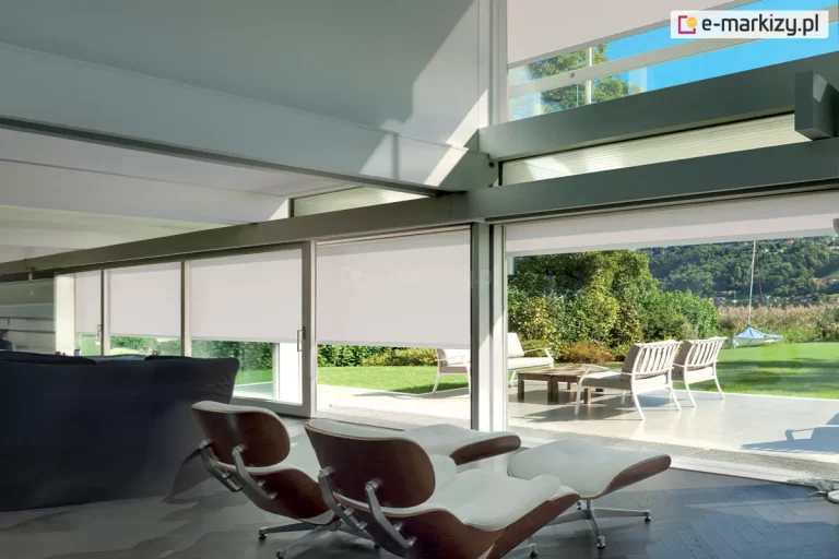 Stylowe, przestronne wnętrze salonu z nowoczesnym designem, którego głównym elementem są duże przesuwne okna wychodzące na taras z widokiem na ogród i jezioro. W oknach zamontowane zostały subtelne, białe rolety screenowe Refleksol 103 Selt, które harmonijnie wpisują się w architekturę wnętrza, podkreślając jego lekkość i nowoczesność. We wnętrzu widoczne są dwa designerskie fotele typu lounge z podnóżkami, tapicerowane białą skórą i drewnem giętym, skierowane w stronę panoramicznego widoku za szybą. Poza przeszkloną ścianą znajduje się taras z białymi fotelami i drewnianym stolikiem kawowym.